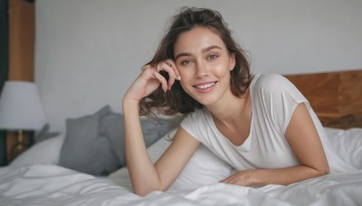 1girl,solo,long hair,looking at viewer,smile,open mouth,blue eyes,brown hair,shirt,white shirt,short sleeves,lying,teeth,indoors,grin,blurry,lips,fingernails,pillow,bed,depth of field,blurry background,on bed,on stomach,t-shirt,messy hair,freckles,realistic,blanket,lamp,ring