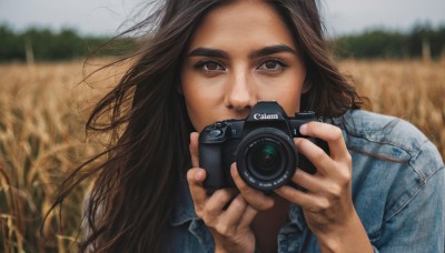 1girl,solo,long hair,looking at viewer,brown hair,shirt,holding,brown eyes,jacket,upper body,outdoors,day,blurry,depth of field,blurry background,thick eyebrows,denim,wind,freckles,jeans,realistic,camera,field,holding camera,denim jacket