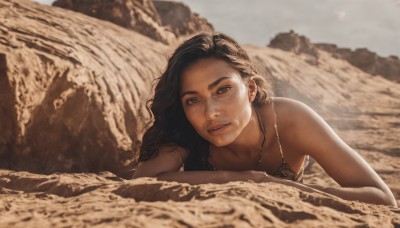 1girl,solo,long hair,looking at viewer,smile,brown hair,black hair,bare shoulders,brown eyes,jewelry,upper body,outdoors,lying,parted lips,dark skin,necklace,blurry,dark-skinned female,lips,blurry background,wavy hair,crossed arms,on stomach,freckles,realistic,nose,sand,sky,day,depth of field,thick eyebrows,rock,sepia,dirty,tribal