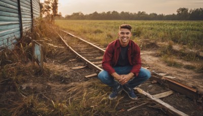 solo,looking at viewer,smile,short hair,shirt,black hair,1boy,holding,sitting,jacket,male focus,outdoors,open clothes,shoes,teeth,pants,black footwear,grin,open jacket,tree,black shirt,facial hair,grass,denim,sneakers,scenery,red jacket,jeans,blue pants,very short hair,undercut,fishing rod,fishing,brown hair,full body,boots,sky,day,squatting,own hands together,sunset,blue footwear,realistic,indian style,field,photo background,railroad tracks